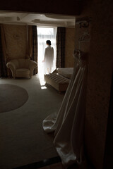 room with soft lighting. In the center of the room stands a woman in a white robe, looking out the window through which light comes in. 