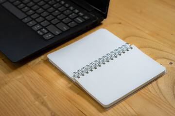 An open spiral notebook with blank pages placed on a wooden desk beside a laptop. Ideal for concepts of work, creativity, or study in a minimalist workspace setting.