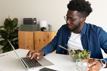 Young black man eating healthy salad while using laptop at home office.