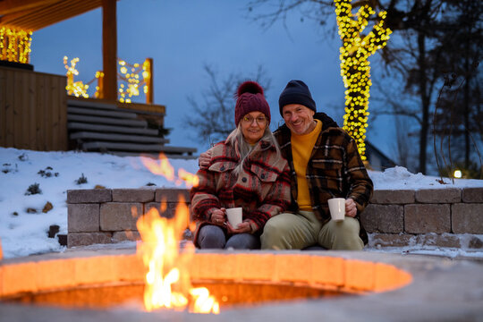 Older couple has lit a fire in garden fire pit. Seniors watching flames, relaxing and drinking hot tea.