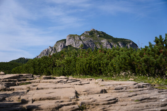 Rocky Hiking Path Surrounded By Small Green Bushes, A Large Cliff Rises In The Background, Blue Cloudy Sky  In The Ceahlău Massif (Masivul Ceahlău)