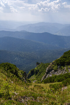 Rolling Hills And Distant Mountains Stretch Into The Horizon, Framed By Cliffs And Greenery Beneath A Cloudy Sky  In The Ceahlău Massif (Masivul Ceahlău)