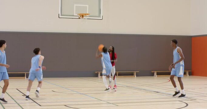 Multiracial male basketball players throwing ball into basket with coach in gym