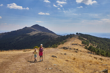 Girl and woman from behind hiking along the mountain ridge towards a peak on the distant horizon,...
