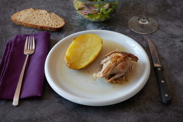 White plate with roasted lamb and baked potato, fork, knife, purple cloth napkin, green lettuce salad, slice of bread and wine cup on a marble texture table