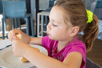 side of four years old blonde little girl with pink shirt and pigtail eating bread with hands, sitting indoor in restaurant