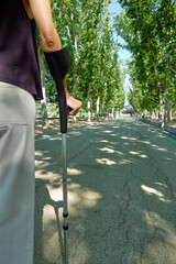 Vertical shot of woman's hand holding or leaning on a crutch on the sidewalk in a park with trees outdoors in a city