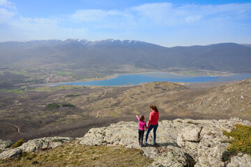 Woman and girl on top of a mountain overlooking the Lozoya valley. Daughter pointing her finger at the Pinilla reservoir and the mountain range of the Guadarrama National Park, in Madrid, Spain