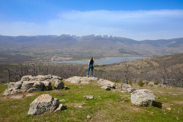 Woman standing with her back on a rock at the top of a mountain looking at the Lozoya Valley reservoir, with Pinilla village and the mountains of the Guadarrama National Park, in Madrid, Spain