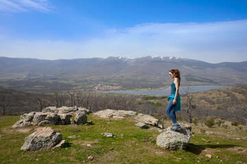 Woman walking on a rock at the top of a mountain above the Lozoya Valley reservoir, with Pinilla village and the mountains of the Guadarrama National Park in the background, in Madrid, Spain