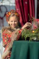 A blonde girl in a national Russian costume on the Maslenitsa holiday. A child in a traditional costume with fur trim and a headdress against a festive backdrop. Winter scene with a beautiful girl