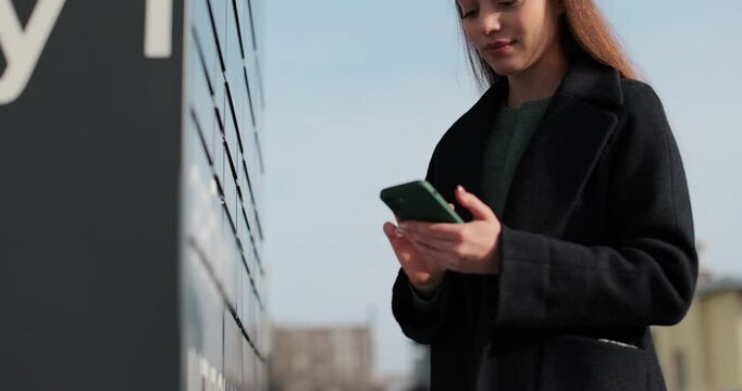 Woman receiving parcel from post terminal machine using smartphone outdoors. Parcel delivery machine. Mail delivery and post service, online shopping, e commerce concept.