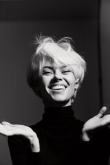 Joyful young woman with short blonde hair expressing excitement, wearing a black turtleneck,...