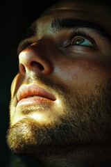 Obraz premium A handsome man, a close-up of his face in the dark, illuminated by sunlight from above. The light shines on his eyes and nose, creating a soft glow effect. He is looking up at something