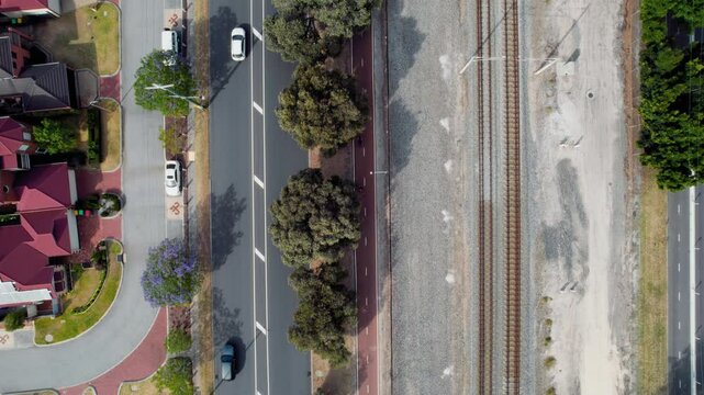 Aerial view of Cars travel down a road parallel to railway tracks with train on it near residential houses and greenery in a suburban setting.