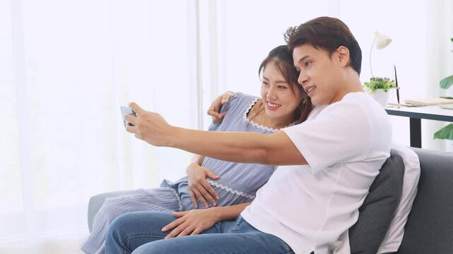 Expectant mother healthy takes selfie with her husband on a smartphone as a keepsake before giving birth. The two look cheerful and happy together sitting on a sofa at home.