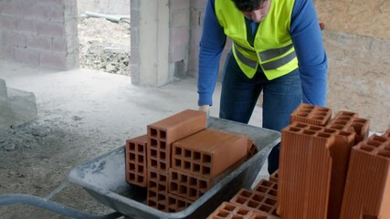 4K slow-motion footage of a construction worker in a safety vest and gloves placing bricks into a partially filled wheelbarrow at a construction site, showcasing effort and skill.