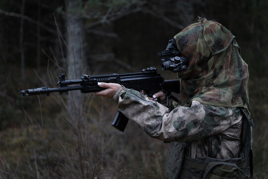 A young girl special forces soldier in a gas mask, camouflage and a tactical helmet with a night vision monocular in the forest at dusk.