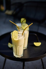 Glasses of lime and condensed milk cocktails sit on a black table in a sunlit terrace. Fresh lime wedges garnish each drink, highlighting a casual summer vibe.
