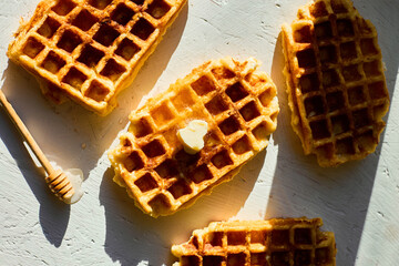 Waffles are arranged on a light surface, with one piece showing a small pat of butter and a honey...