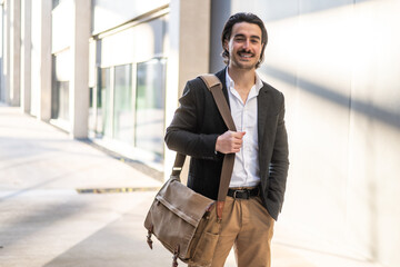 Fototapeta premium Young businessman smiling while walking outside a modern office building, carrying a messenger bag and enjoying a sunny day on the way to work, exuding confidence and positivity