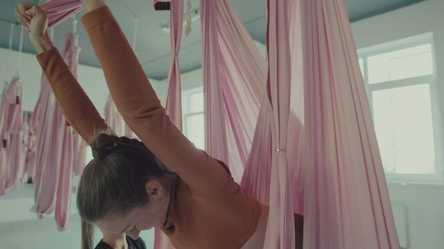 Women stretch and perform inversion poses in a spacious studio featuring flowing pink hammock for aerial yoga.