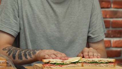 Man preparing lunch. Man sitting at table at home in the kitchen and preparing sandwich snack, demonstrating prepared sandwich cutting in half on the board.