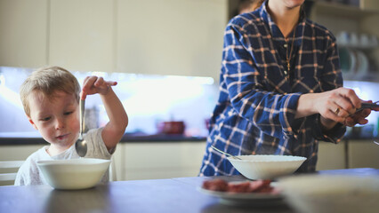 Mother warming hearty soup while young son eagerly eating at rustic kitchen table