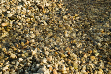 Pebbles and stones beneath the shallow surface of crystal-clear water. The sunlit scene creates...