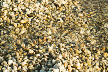 Pebbles and stones beneath the shallow surface of crystal-clear water. The sunlit scene creates shimmering reflections highlighting the textures and colors of the stones.