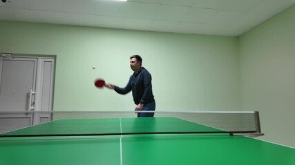 Casual male player showing table tennis stance and techniques at green ping-pong table, captured in well-lit mint-colored room with professional equipment setup.