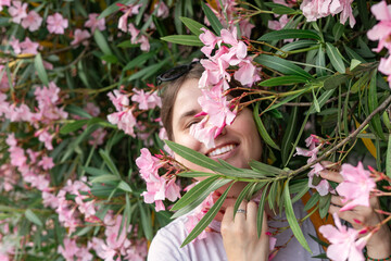 cheerful woman surrounded by vibrant pink oleander flowers, smiling warmly while playfully engaging...