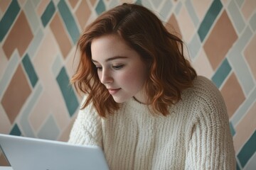Young redhead woman using laptop, focused on screen, sitting against a geometric patterned wall.