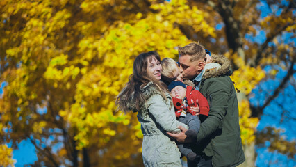 Fototapeta premium Parents holding their children in a park, surrounded by beautiful fall foliage and enjoying a sunny day