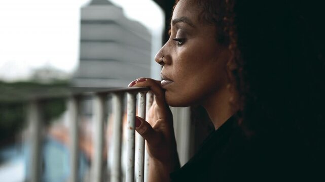 Profile close-up of a serious black woman leaning ln metal bar by home balcony gazing downwards with melancholic expression struggling but observing surroundings
