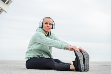 Fototapeta premium Mature Woman Stretching Her Legs While Listening to Music with Headphones on, Embracing a Sporty Lifestyle and Prioritizing Fitness and Well-being Through Regular Exercise and Stretching Routines