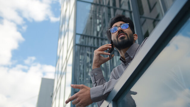 Bottom view Arabian businessman speak mobile phone outdoors on terrace serious Indian man talk with business partner smartphone call negotiate solve male CEO employer talking cellphone on city balcony