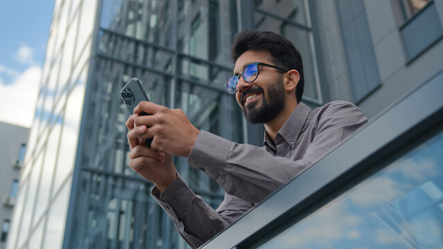 Smiling Arabian Indian man businessman entrepreneur manager use mobile phone application in city office terrace bottom view male business owner employer browsing social media texting cellphone message