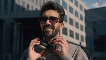 Carefree Arabian Indian man in sunglasses wear headphones listen music in sunny city joyful happy businessman guy listening song outdoors enjoy sound in earphones near business office company outside
