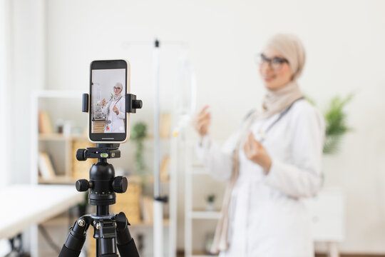 Young Muslim woman in medical attire records video tutorial about intravenous dropper setup using smartphone. Educational content creation in a healthcare environment with modern technology.