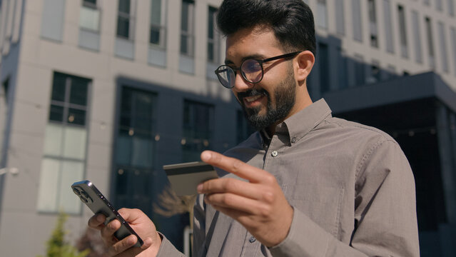 Arabian Indian man businessman hold bank credit card pay purchases with mobile phone outdoors male customer client enter security code buying paying for service business payment money transfer in city
