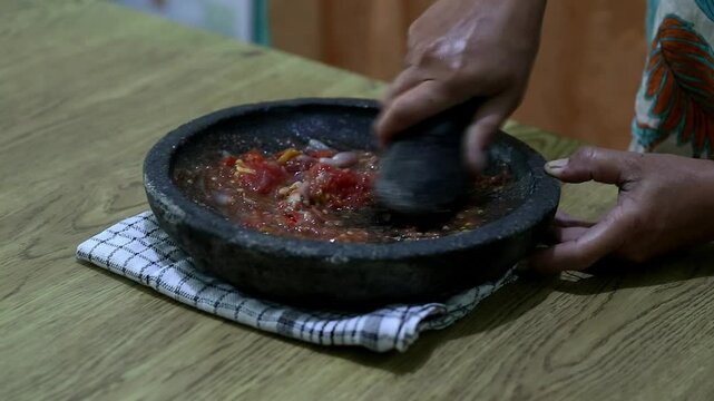 Indonesian woman's hands grinding spices on a stone mortar for sambal. Traditional Indonesian sambal being prepared with mortar and pestle. Asian womans hands crushing chili peppers and spices 
