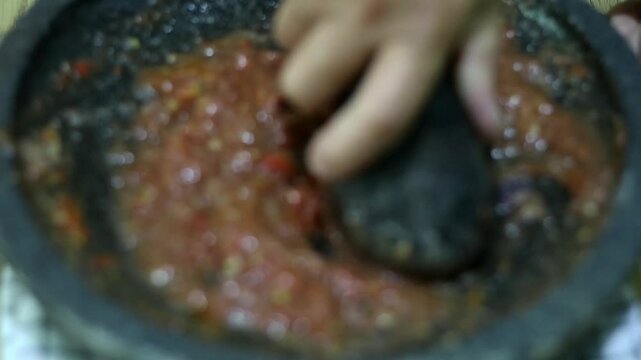 Indonesian woman's hands grinding spices on a stone mortar for sambal. Traditional Indonesian sambal being prepared with mortar and pestle. Asian womans hands crushing chili peppers and spices 