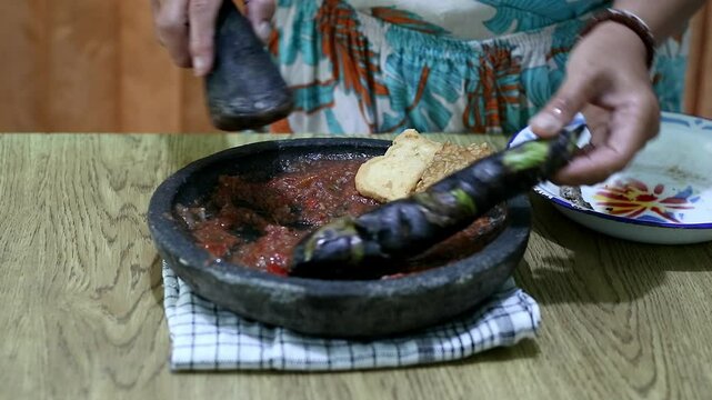 Indonesian woman's hands grinding spices on a stone mortar for sambal. Traditional Indonesian sambal being prepared with mortar and pestle. Asian womans hands crushing chili peppers and spices 