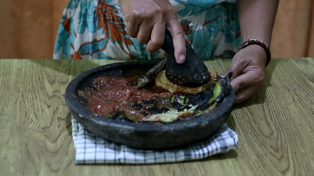 Indonesian woman's hands grinding spices on a stone mortar for sambal. Traditional Indonesian sambal being prepared with mortar and pestle. Asian womans hands crushing chili peppers and spices 