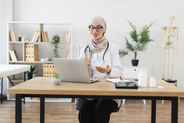 Muslim female doctor having video call with patient using laptop in modern office setting. Middle-Eastern woman wearing hijab and lab coat, discussing healthcare solutions through online communication