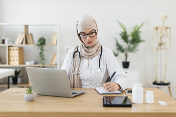 Young Muslim female doctor wearing hijab conducting video call with patient, writing prescription....