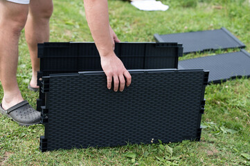 Close-up of hands assembling dark gray plastic panels on green grass. The textured panels appear...