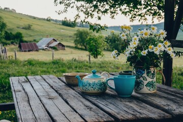 Tea in the Countryside: A rustic countryside setting with a wooden table, a pot of tea, and handmade ceramic cups, surrounded by rolling hills and farms.