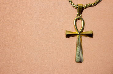 The image shows a gold pendant in the shape of an ankh hanging on a chain. The ankh is an ancient Egyptian symbol representing life and immortality. The background of the image is light beige.
